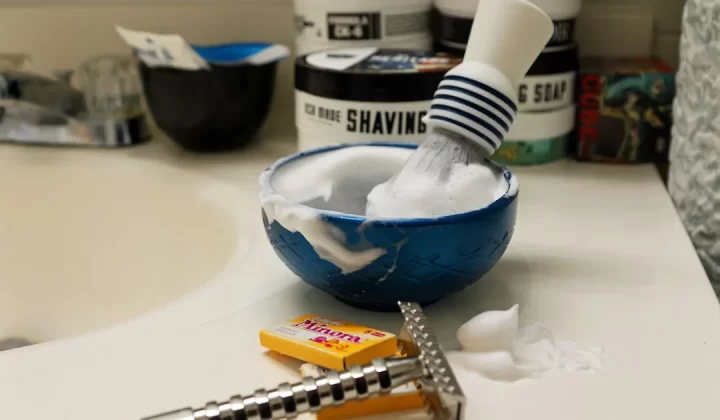 Shaving brush and bowl with lather and razor on sink.